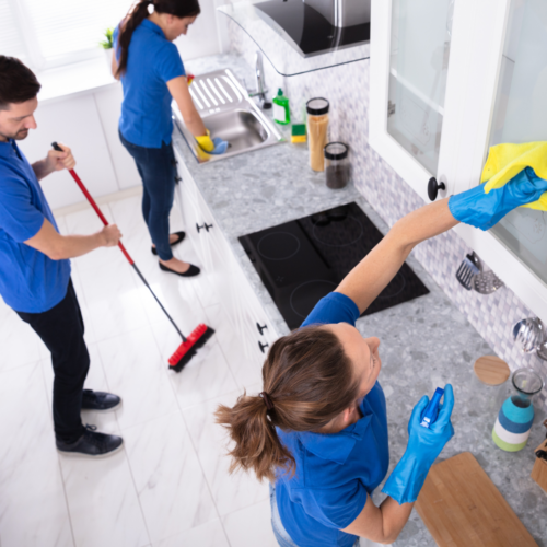 Team of professional cleaners in blue uniforms thoroughly cleaning a modern kitchen, one person wiping cabinets, another scrubbing the floor, and a third washing dishes, showcasing Clean Corps' residential cleaning services.