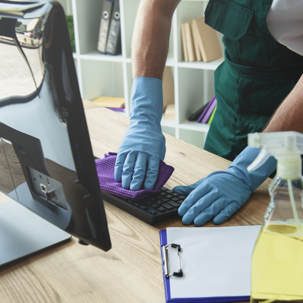 Commercial cleaning professional wearing blue gloves, cleaning a keyboard with a purple cloth, alongside a spray bottle and clipboard, in a tidy office environment.