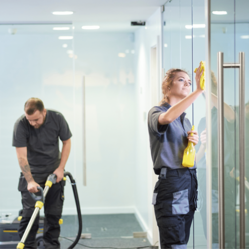 Cleaning professionals performing post-construction cleaning, one using a vacuum and the other cleaning glass surfaces with a spray bottle, in a modern office setting.