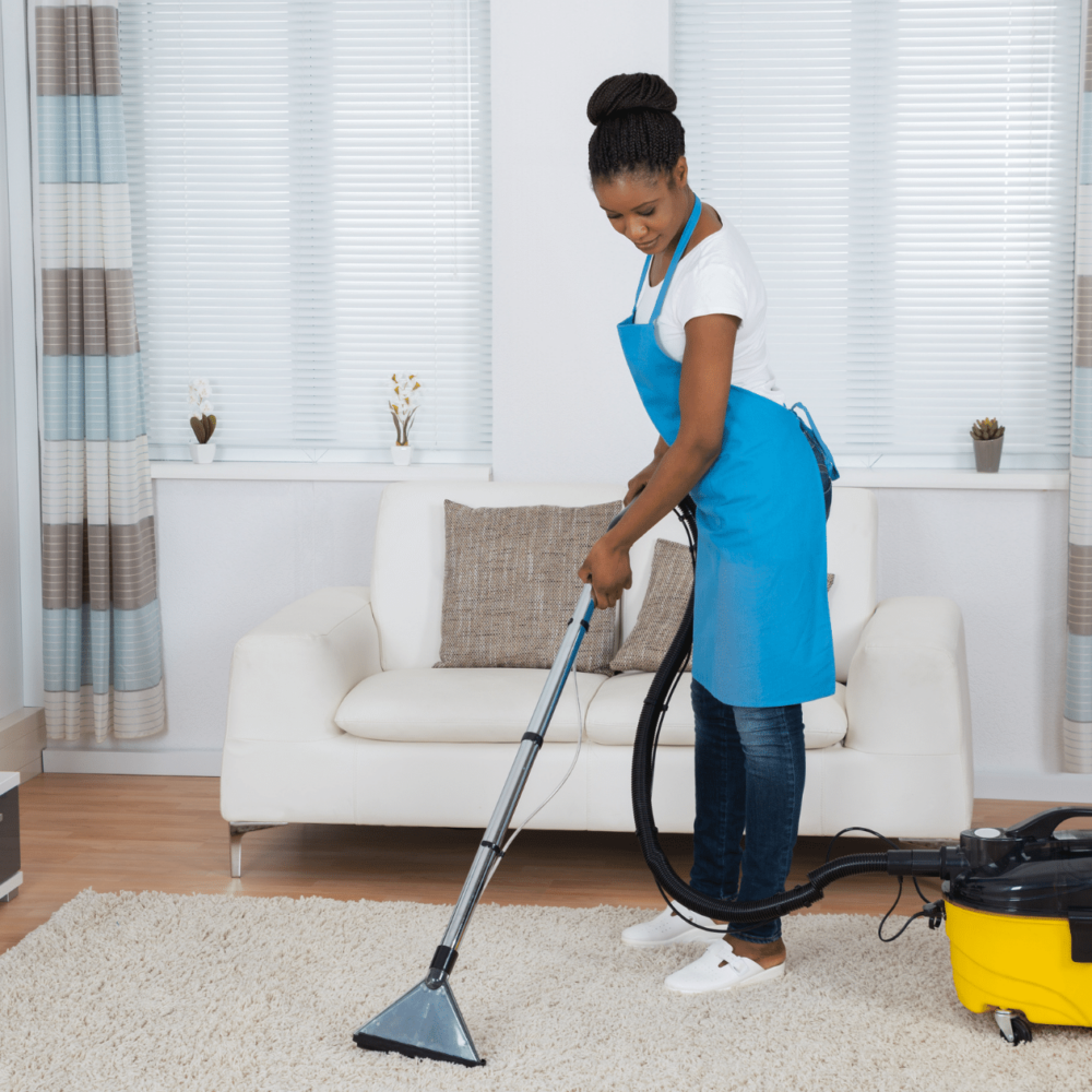 Woman in blue apron using a carpet cleaning machine on a beige rug in a modern living room, showcasing residential cleaning services by Clean Corps.