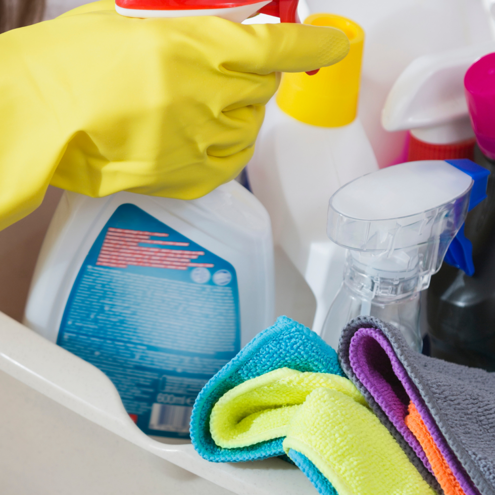Hand in yellow rubber glove holding a spray bottle, surrounded by various cleaning solutions and colorful microfiber cloths, representing cleaning services offered by Clean Corps.