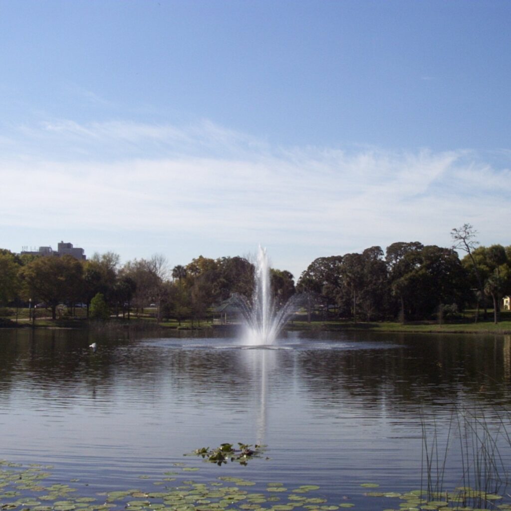 Fountain on Lake Lily with lily pads and oak-lined shoreline under a blue sky in Maitland, Florida
